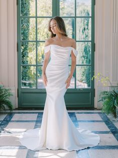 a woman standing in front of a door wearing a white wedding dress with off the shoulder sleeves