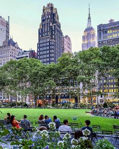 many people are sitting on benches in the park with tall buildings and trees behind them