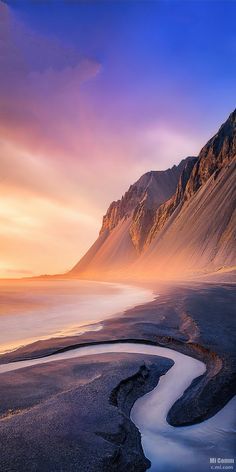 a river running through a valley next to a mountain range at sunset with the sun going down