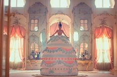 a woman standing on top of a large cake in a room filled with windows and curtains