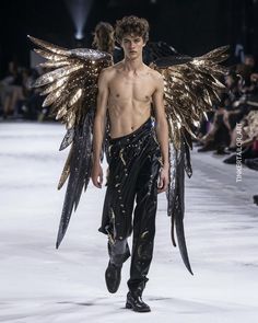 a man walking down a runway with gold wings on his chest and black leather pants