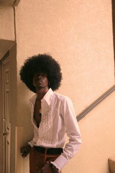 a woman with an afro standing in front of a wall wearing a white shirt and brown pants