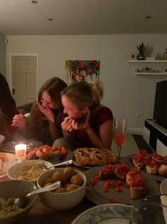 three women sitting at a table with food and drinks in front of them, while one woman holds her face to her mouth