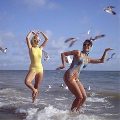 two women in swimsuits run into the ocean while seagulls fly overhead