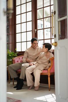 an older man and young woman sitting on a pink chair in front of large windows