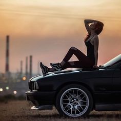 a woman sitting on the hood of a black car in front of an industrial area