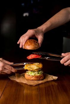 two hands holding knives over a hamburger on top of a wooden table