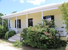 a white house with black shutters and flowers on the front porch, surrounded by shrubs