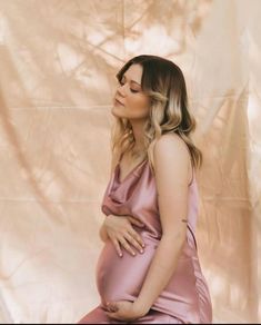 a pregnant woman in a pink dress sitting on a white sheet with her hands under her stomach