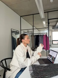 a woman sitting at a table with a laptop computer in front of her and clothes on racks behind her