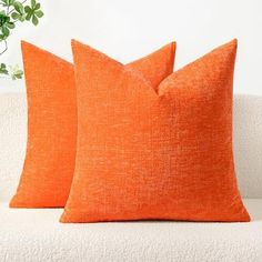 two orange pillows sitting on top of a white couch next to a potted plant