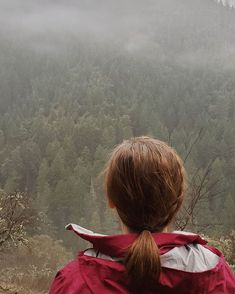 a woman in a red jacket looking out over a forest on a foggy day