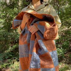 a woman is wrapped in an orange and grey blanket while standing outside with trees behind her