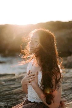 a woman with long hair standing on the beach at sunset, looking into the distance