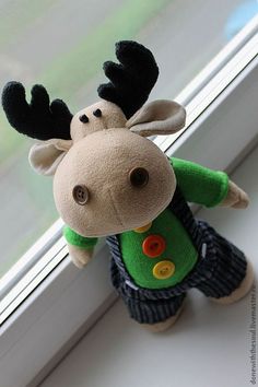 a stuffed moose wearing a green shirt and blue pants sitting in front of a window