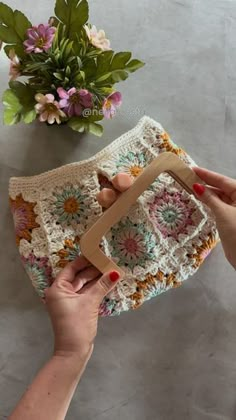 two hands holding a crocheted purse next to a potted plant on a table