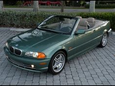 a green convertible car parked in front of a brick sidewalk with trees and bushes behind it