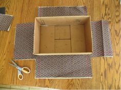 an open cardboard box sitting on top of a wooden floor next to scissors and tape