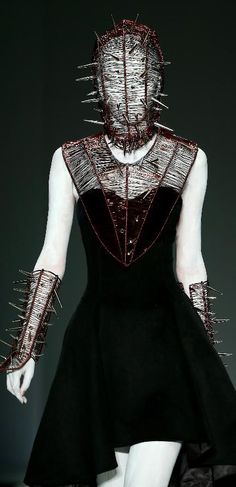 a woman in a black dress with lots of spikes on her head and arms, walking down a runway