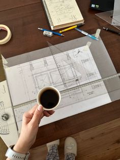 a person holding a cup of coffee on top of a wooden table next to some construction drawings