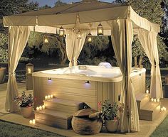 an outdoor hot tub with lights on the sides and curtains over it, surrounded by greenery