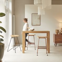 a woman standing at a table with two stools in front of her and a plant on the other side