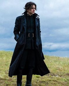 a man standing on top of a grass covered field wearing a black coat and boots