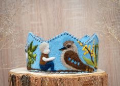 a blue felt crown with a brown bird on it sitting on a tree stump in front of a wooden wall