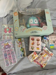 an assortment of toys and stickers in a box on top of a bed with sheets