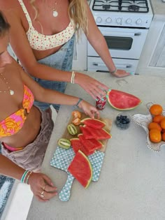 two women in the kitchen slicing up watermelon slices on a cutting board with fruit nearby