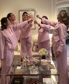 four women in pink pajamas toasting with wine glasses on a coffee table while another woman looks on