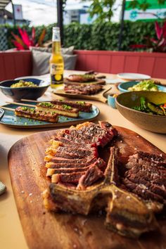 a wooden cutting board topped with sliced up meat and vegetables next to bowls of salad