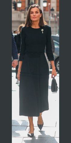 a woman in a black dress walking down the street with her hand on her hip