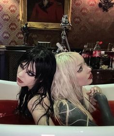 two women sitting in a bathtub with red liquid on the floor and one wearing black hair