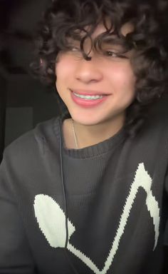 a young man with curly hair wearing a black sweater and headphones smiling at the camera