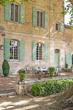 an old stone house with green shutters and tables in the front yard, surrounded by greenery