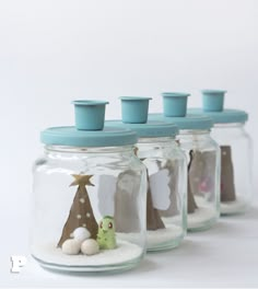 four glass jars filled with sand and small trees in the bottom one has a blue lid