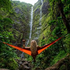 a woman is sitting in a hammock near a waterfall and holding her hands up