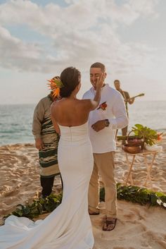 a man and woman standing on top of a sandy beach next to the ocean with an orange flower in their hair