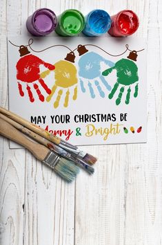 a christmas card with three handprints and paintbrushes next to it on a white wooden surface