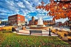 a fountain in the middle of a park surrounded by buildings and trees with fall foliage