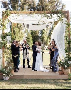 a group of people standing around each other under a white cloth covered arbor with flowers and greenery