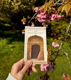 a person holding up an old book in front of some pink flowers on a tree