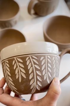 a hand holding a brown and white coffee cup with leaves on the inside, in front of other cups