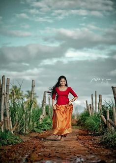 a woman walking down a dirt road in front of tall grass and bamboo poles with clouds overhead