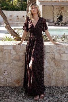 a woman standing next to a stone wall wearing a long dress with a slit down the middle