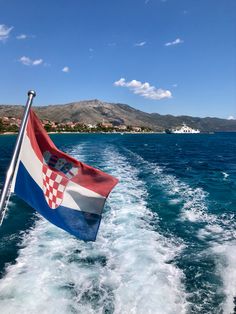 the croatia flag is flying from the back of a boat in the water with mountains in the background