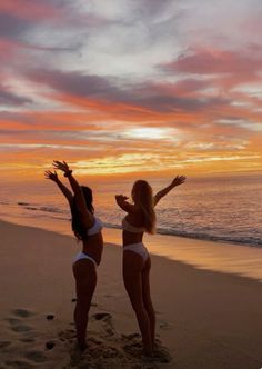 two women in bikinis standing on the beach with their arms up to the sky