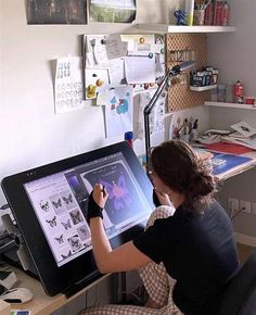 a woman sitting in front of a desk with a drawing tablet on it and papers all over the wall