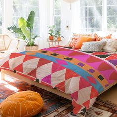 a bed with colorful comforters and pillows in a room next to a plant on the floor